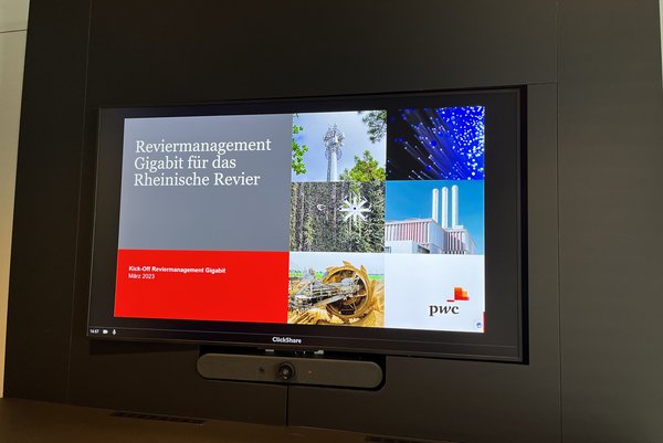 Titelfolie einer Präsentation auf einem Bildschirm in schwarzer Umrahmung mit dem Titel „Reviermanagement Gigabit für das Rheinische Revier“ mit Symbolbildern eines Funkturms, einer Glasfasernahaufnahme, einer Drohne beim Einsatz in der Landwirtschaft, einem Industriegebäude mit 3 Schlöten und einem Braunkohlebagger. In der rechten unteren Ecke ist das Logo der PricewaterhouseCoopers Wirtschaftsprüfungsgesellschaft abgebildet. Unter dem Bildschirm befindet sich eine Webcam.